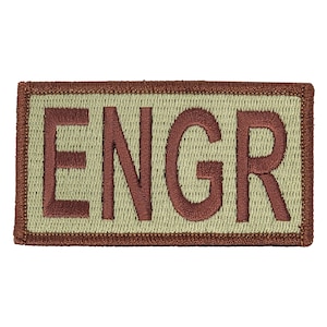 Może przedstawiać: Prostokątny, beżowy naszywka z brązową haftowaną ramką i napisem "ENGR" brązowymi literami.