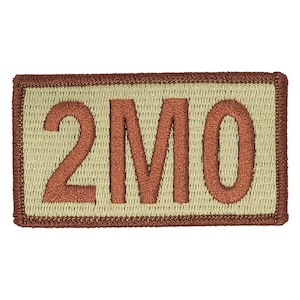 May include: A tan-colored rectangular patch with a brown embroidered border and the text "2MO" in brown.