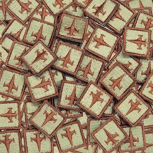 May include: A close-up of a pile of brown and green fabric patches with an embroidered image of a fighter jet on each patch.