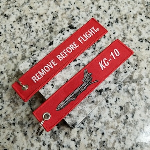 May include: Two red fabric keychains. One keychain has the text "REMOVE BEFORE FLIGHT" in white. The other keychain has the text "KC-10" in white, with a detailed illustration of an aircraft. Both keychains have a silver metal ring.