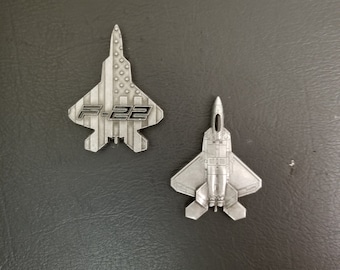 May include: Two silver metal F-22 fighter jet pins. One pin features a US flag design with the text "F-22" in black. The other pin is a solid silver design.