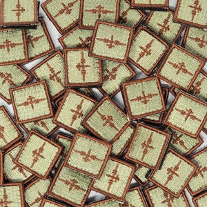 Puede incluir: Primer plano de una colección de pequeños parches cuadrados con un borde marrón y un fondo verde. Cada parche presenta un diseño estilizado bordado de una cruz con múltiples líneas que se extienden desde cada brazo.