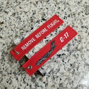May include: Red fabric keychain with white text that reads "REMOVE BEFORE FLIGHT" and a red fabric keychain with white text that reads "C-17" and a black silhouette of a plane.