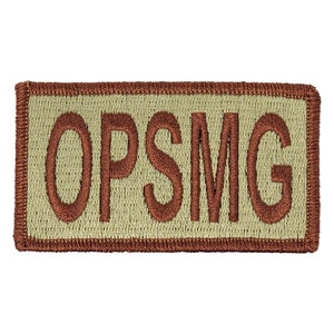 Puede incluir: Un parche de tela beige con un borde bordado marrón y el texto "OPSMG" en hilo marrón.
