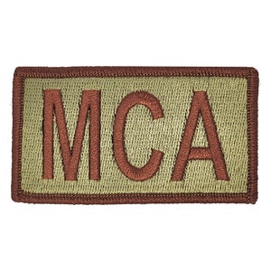May include: A tan rectangular patch with a brown embroidered border and the word "MCA" in brown lettering.