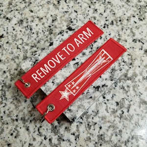 May include: Two red fabric keychains with white embroidery. One keychain says "REMOVE TO ARM" and the other has a star and a stylized jet.