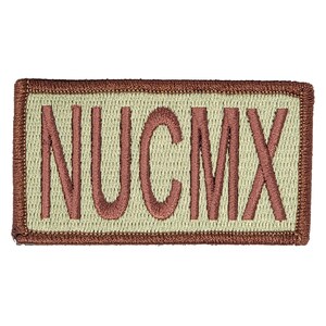Könnte beinhalten: Ein beigefarbener gestickter Aufnäher mit braunem Rand. Der Aufnäher trägt die Aufschrift "NUCMX" in braunem Garn.