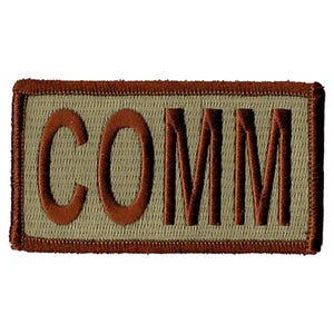 Op de afbeelding: Een rechthoekige patch met het woord "COMM" geborduurd in donkeroranje draad. De achtergrond is lichtbeige en de rand is donkerder bruin. De patch heeft een getextureerd uiterlijk.