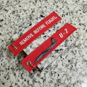 May include: Two red fabric keychains with white stitching. One keychain has the text "REMOVE BEFORE FLIGHT" and the other has the text "U-2" with a black and white illustration of a plane.