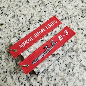 May include: Red fabric keychain with white text that reads "REMOVE BEFORE FLIGHT" and a red fabric keychain with white text that reads "E-3" and a white outline of an airplane.