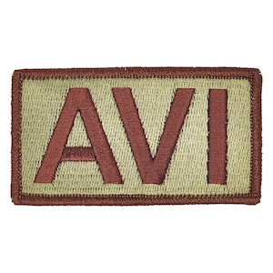 May include: A tan rectangular patch with a brown embroidered border and the word "AVI" in brown letters.