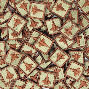 May include: A close-up of a pile of green and brown fabric patches featuring a stylized image of a fighter jet. The patches are arranged in a random pattern.