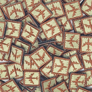 Op de afbeelding: Een close-up van een stapel groene en bruine stoffen patches met een gestileerd vliegtuigontwerp. De patches zijn vierkant en hebben een bruine rand.