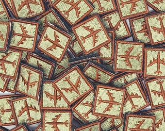 May include: A close-up of a pile of green and brown fabric patches featuring a stylized airplane design. The patches are square and have a brown border.