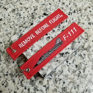 May include: Two red fabric keychains with white embroidery. One keychain says "REMOVE BEFORE FLIGHT" and the other keychain says "F-111" with a small gray airplane graphic.