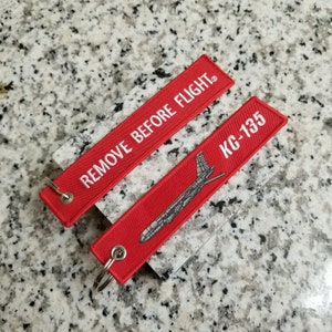 May include: Two red fabric keychains with white stitching. One keychain has the text "REMOVE BEFORE FLIGHT" and the other has the text "KC-135" and a silhouette of an airplane.