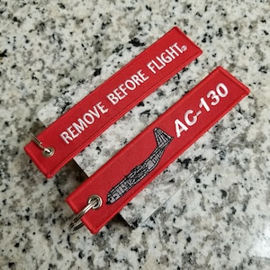 May include: Two red fabric keychains with white embroidery. One keychain says "REMOVE BEFORE FLIGHT" and the other says "AC-130" with a silhouette of an airplane.