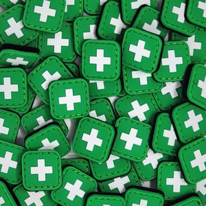 May include: A close-up of a pile of green and white square patches with a white plus sign in the center. The patches have a white stitched border.