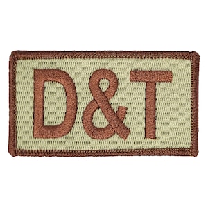Könnte beinhalten: Ein braunes gesticktes Patch mit den Buchstaben "D&T" auf beigefarbenem Hintergrund.