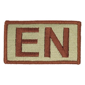 Peut inclure: Un patch rectangulaire beige avec une bordure brodée marron. Les lettres "EN" sont brodées en marron sur le patch.