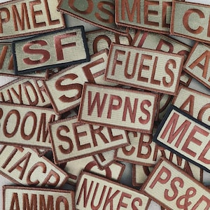 Könnte beinhalten: Eine Ansammlung von gestickten Aufnähern mit verschiedenen Texten, darunter "SF", "FUELS" und "WPN". Die Aufnäher haben einen beigen Hintergrund mit braunen Buchstaben und Rändern. Einige Aufnäher haben einen schwarzen Rand. Die Aufnäher sind verstreut.