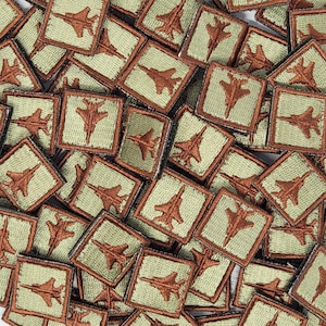 May include: A collection of brown and green fabric patches featuring an embroidered image of a fighter jet. The patches are square shaped and have a light green background with a brown outline.