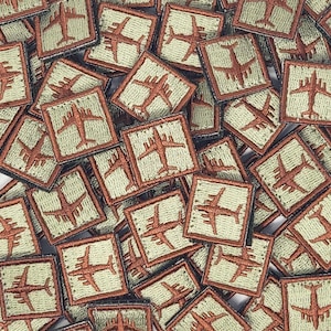 Puede incluir: Primer plano de un montón de parches pequeños y cuadrados. Cada parche está hecho de una tela verde claro con un contorno marrón. Los parches presentan una imagen estilizada de un avión en hilo marrón.