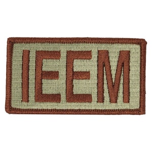 Puede incluir: Un parche bordado marrón con las letras "IEEM" sobre un fondo beige.