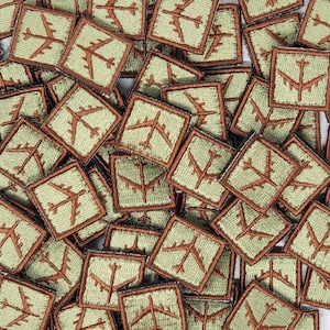 Puede incluir: Primer plano de una pila de parches de tela marrón y verde con una imagen bordada de un avión. Los parches son todos del mismo tamaño y forma, y están dispuestos en un patrón aleatorio.