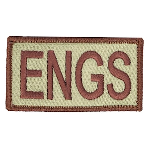 Può includere: Un cerotto rettangolare beige con un bordo ricamato marrone e la parola "ENGS" in lettere marroni.