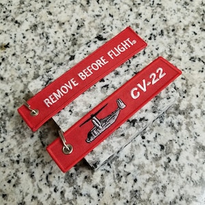 May include: Two red embroidered fabric tags. One tag reads "REMOVE BEFORE FLIGHT" in white. The other tag has a detailed image of a helicopter and the text "CV-22" in white. Both tags have a silver metal ring.