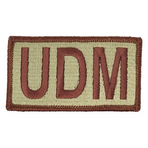 May include: A tan-coloured patch with a brown embroidered border and the text "UDM" in brown.