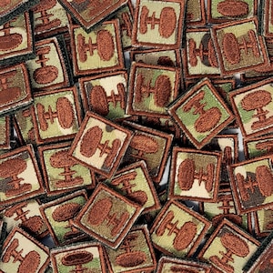 May include: A pile of square patches with a brown and green camouflage design. Each patch has a brown emblem, possibly a wheel or gear. The patches are closely arranged, forming a textured, repetitive pattern.