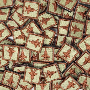 Może przedstawiać: Zbliżenie na stos małych, kwadratowych naszywek z haftowanym brązowym wizerunkiem myśliwca na jasnozielonym tle.