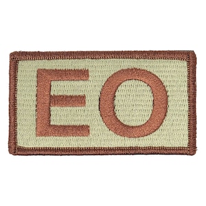 Puede incluir: Un parche rectangular de color beige con un "EO" bordado en marrón en el centro.