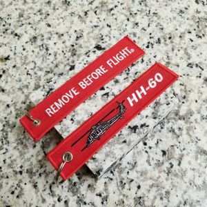 May include: Red fabric keychain with white text that says "REMOVE BEFORE FLIGHT" and a red fabric keychain with white text that says "HH-60" and a black and white image of a helicopter.