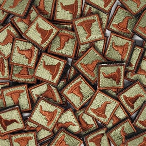 Könnte beinhalten: Ein Stapel kleiner, quadratischer Stoff-Patches in Grün und Braun mit einem gestickten Bild eines Hexenhutes auf jedem Patch.