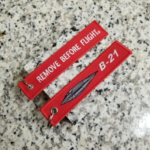 May include: Two red keychains with white text. One keychain says "REMOVE BEFORE FLIGHT" and the other says "B-21" with a gray silhouette of a plane.