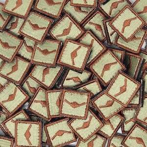 May include: A close-up of a pile of small, square patches. Each patch is made of a light green fabric with a brown, stylized arrow design. The patches are arranged in a random pattern.