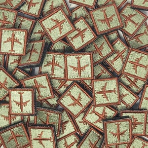 May include: A close-up of a pile of green and brown fabric patches with an embroidered airplane design. The patches are all the same size and shape, and they are arranged in a random pattern.