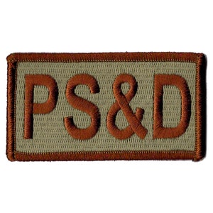 Puede incluir: Parche bordado rectangular con las letras "PS&D" en color óxido. Las letras están cosidas sobre un fondo verde oliva claro, enmarcado por un borde color óxido.