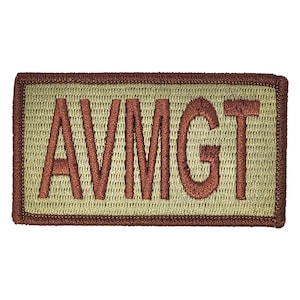 May include: A tan-colored patch with a brown embroidered border and the text "AVMGT" in brown.