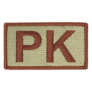 Puede incluir: Parche bordado rectangular con las letras "PK" en marrón oscuro. Las letras están sobre un fondo beige claro, enmarcadas por un borde marrón. Ideal para personalizar prendas y accesorios.
