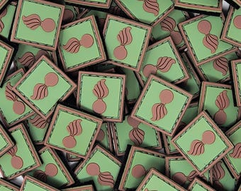 Op de afbeelding: Een verzameling vierkante patches met een groene achtergrond en een bruin embleem. Het embleem heeft een gestileerd ontwerp met een cirkelvormige basis en een vlamachtig element. De patches hebben een bruine rand.