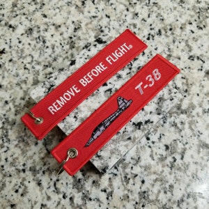 May include: Two red keychains with white text. One keychain says "REMOVE BEFORE FLIGHT" and the other says "T-38" with a small silhouette of a plane.