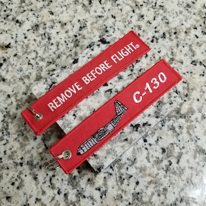 May include: Two red fabric keychains with white embroidery. One keychain says "REMOVE BEFORE FLIGHT" and the other says "C-130" with a small airplane graphic.