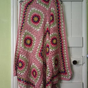 May include: A pink crochet blanket with a floral design. The blanket has circular patterns in pink, green, white, purple, and yellow. It is hanging on a white door with black hooks. The blanket is approximately 150 cm x 150 cm.