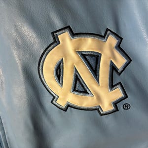 May include: Light blue jacket with a large embroidered logo. The logo is a white and navy blue interlocking "NC" design, with a black outline. The jacket has a zipper and a textured, leather-like appearance.