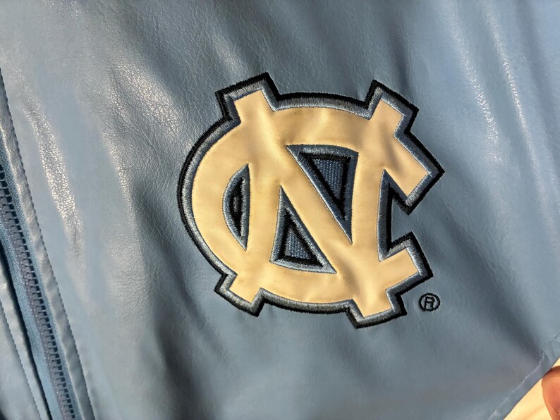 May include: Light blue jacket with a large embroidered logo. The logo is a white and navy blue interlocking "NC" design, with a black outline. The jacket has a zipper and a textured, leather-like appearance.