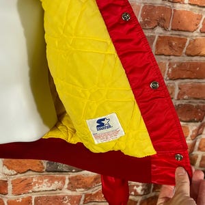 May include: Red and yellow Starter jacket with a quilted yellow lining. The jacket has a red exterior with snap closures and a red collar. The Starter logo is visible on a tag inside the jacket.